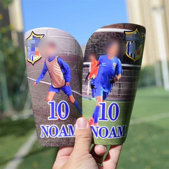 Pair of full-size shin guards printed '10 NOAM' with player photos, held up against the sky