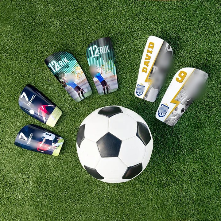 Six printed shin guards with names, numbers and photos arranged around a football on turf